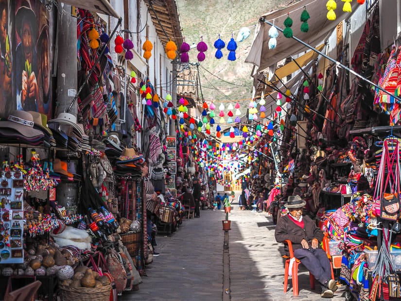 A tourist market street in Pisac with a local man sitting in a chain on the side and colorful decorations hanging above the street
