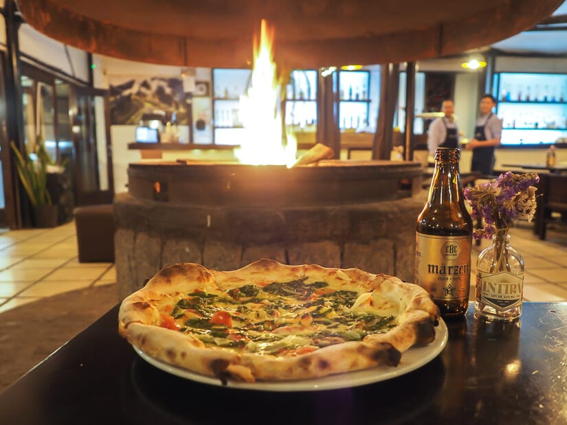 Close up of an Italian-style pizza with a round fireplace in the background and two waiters standing behind it