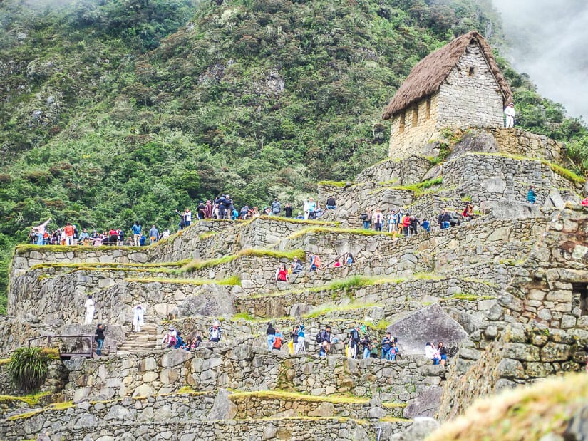 Crowds of people standing on stone terraces below the Guardian House at Machu Picchu