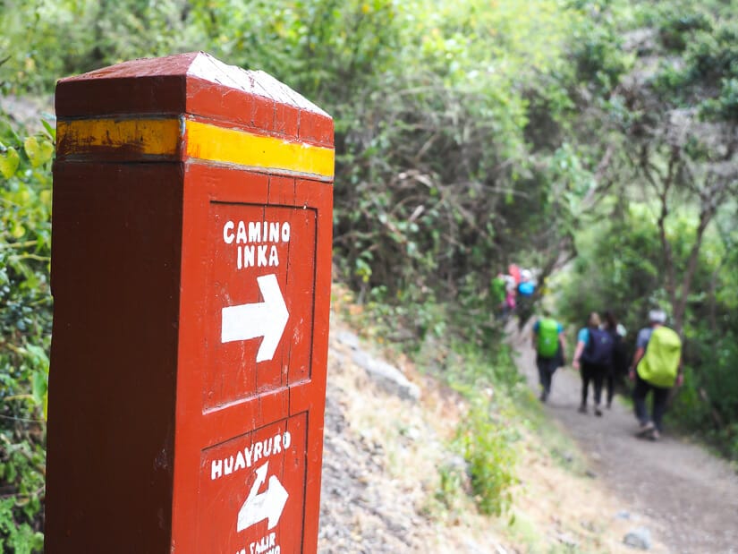 A red wooden sign post that says "Camino Inka" with arrow pointing to the Inca Trail, and some trekkers in the background
