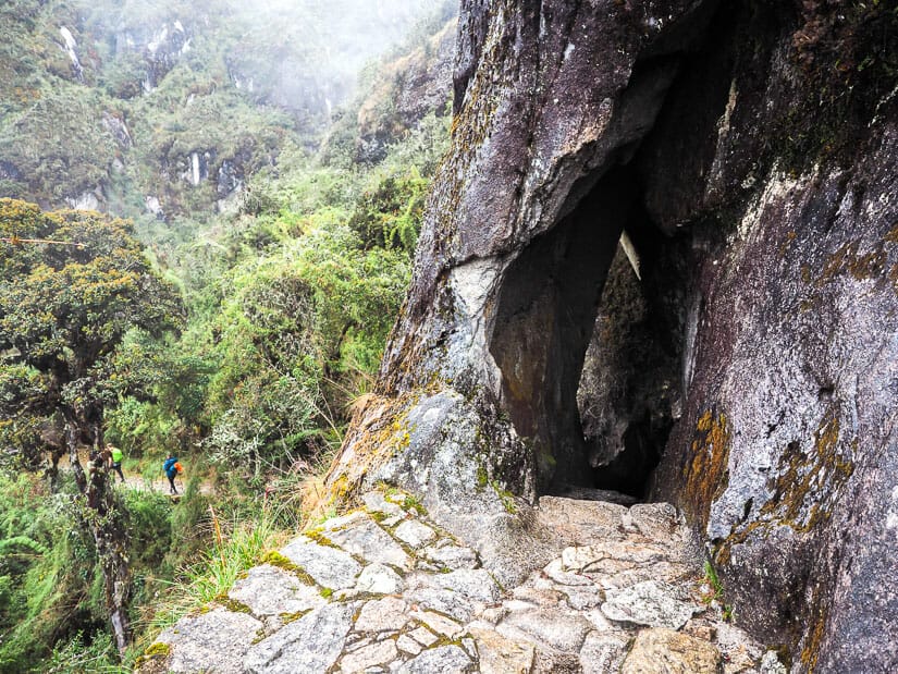 A stone path of the Inca Trail passes through a cave, with some trekkers visible on the path in the distance.