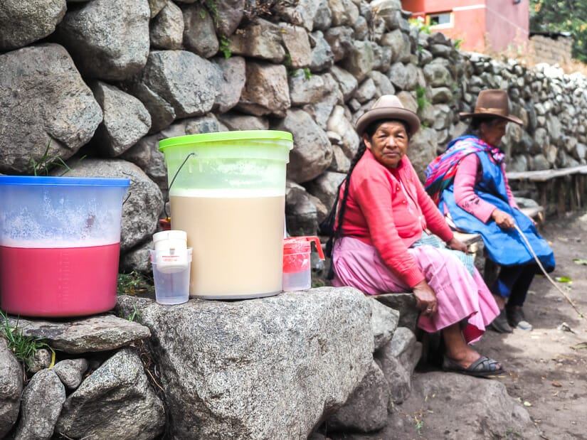 Two Quechua women sitting beside a stone wall on the Inca Trail with a big jug of chicha and other or a pink drink