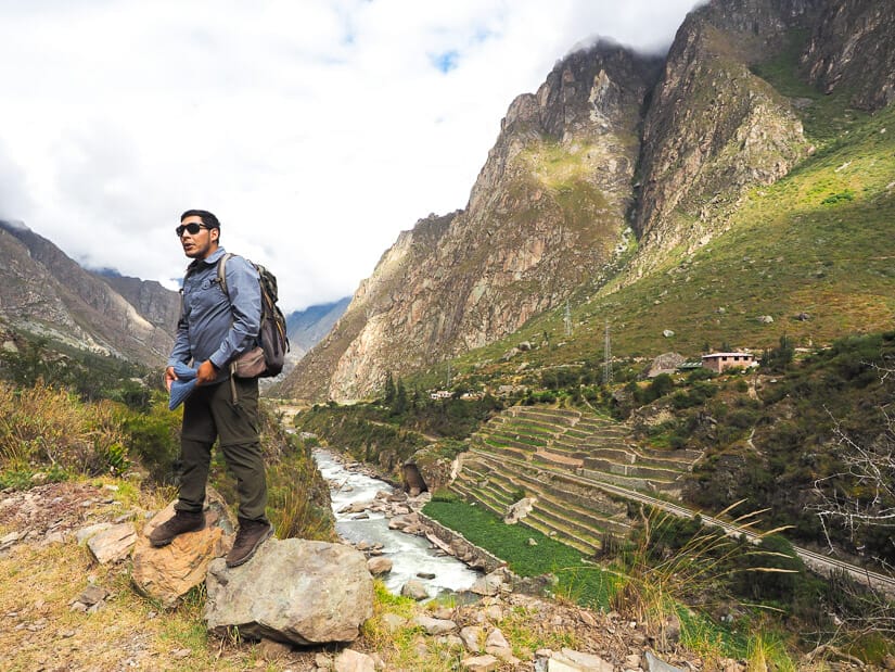 An Inca Trail guide standing on rocks, with a river and some Inca ruins behind him