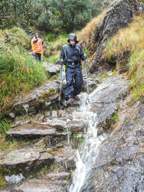 A male trekker hiking down stone stairs with water running down them, with another female trekker behind
