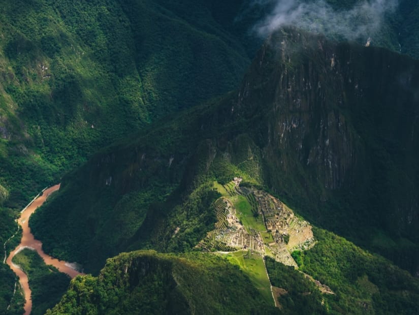 A view of Machu Picchu ruins shot from on top of a mountain way above it, with verdant mountains all around the site and a brown river visible far below to the left