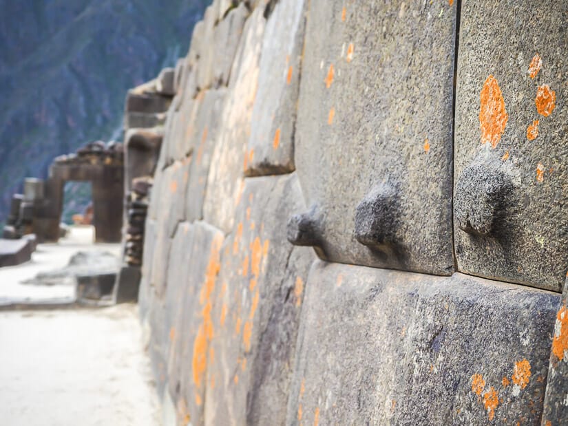 Bumps sticking out from a stone wall with orange spots on it, with Ollantaytambo Sun Gate visible at end of wall
