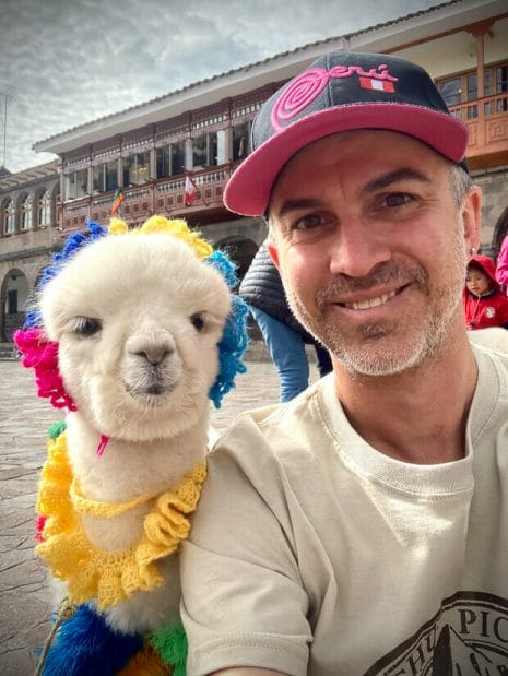 Nick Kembel posing with a baby alpaca wearing colorful accessories in Cusco Peru