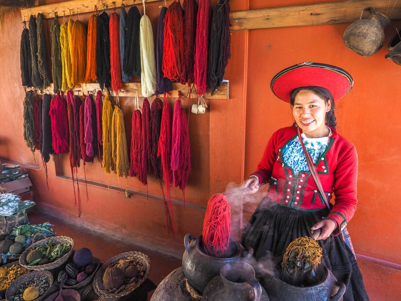 A Quechua woman in colorful clothing lifting up two clumps of steaming dyed yarn, with colorful yarns hanging on the wall behind her
