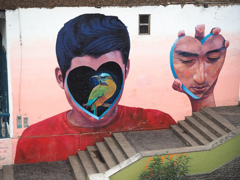 A painted mural of a man with a heart shaped hole in his face with a bird in it and he's holding the face slice in his hand