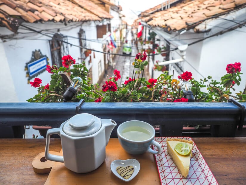 A tea set and lemon pie slice on a table, with flowers in front of it and looking down on a narrow alley in San Blas, Cusco