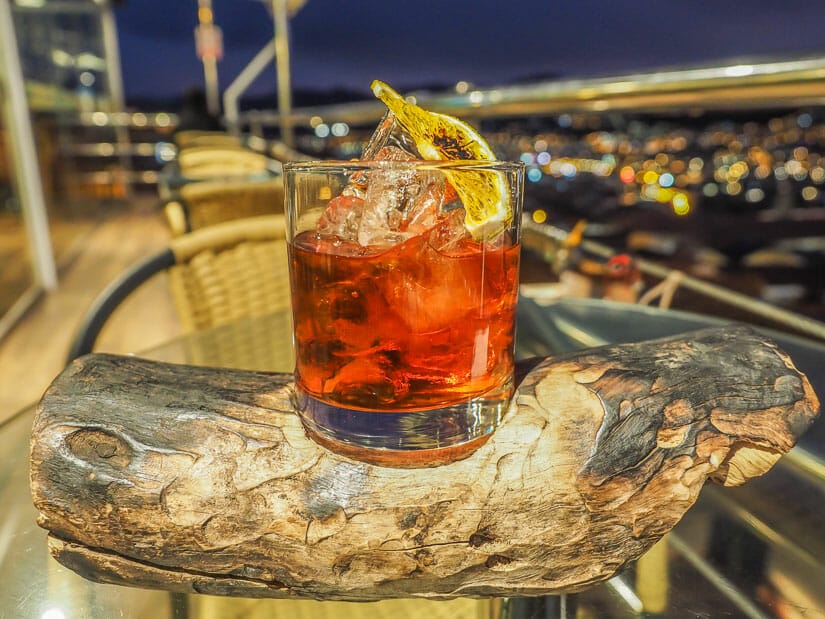 An orange cocktail in a glass on a piece of wood with view of Cusco at night to the side