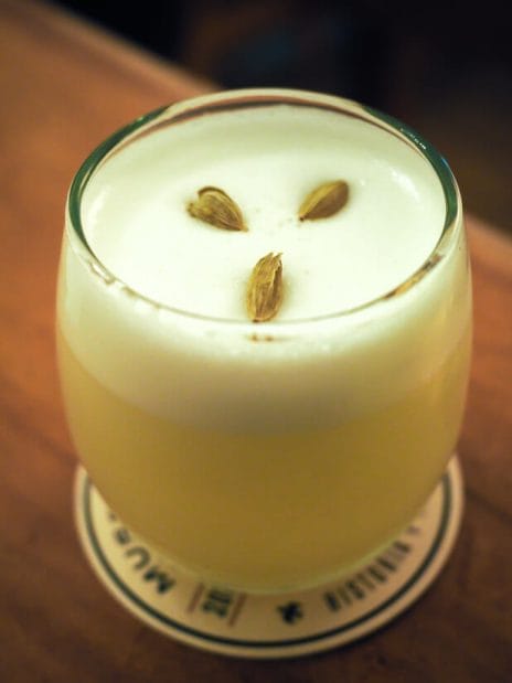 A glass of cardamom infused pisco sour with three cardamom pods on top