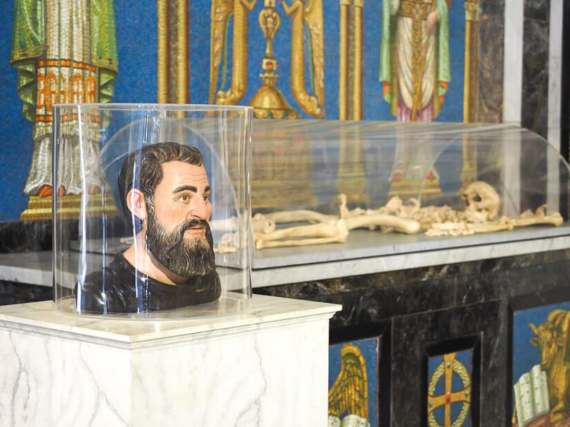 A bust statue of Pizarro and his skeleton in a glass case behind with painted walls
