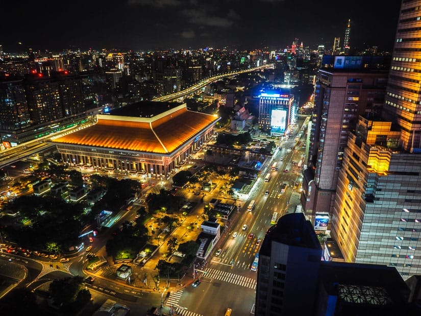 A view looking down at Taipei at night, with large Taipei Train Station building, long road full of cars, and skyscrapers in the distance