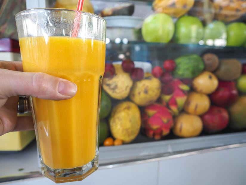 A hand holding a glass or orange colored fresh fruit juice with various fruits behind a glass panel
