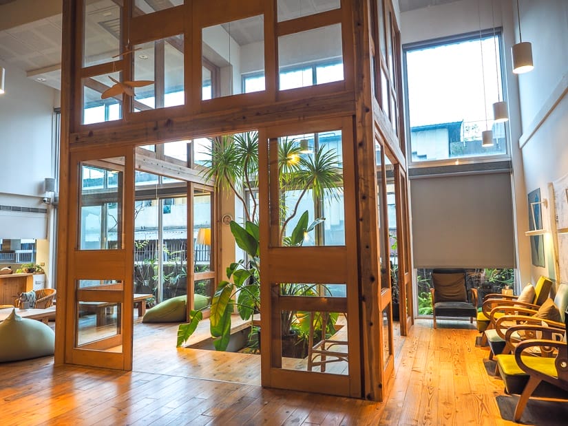 A hostel common room with large windows, plants, and an enclosed wooden sitting room