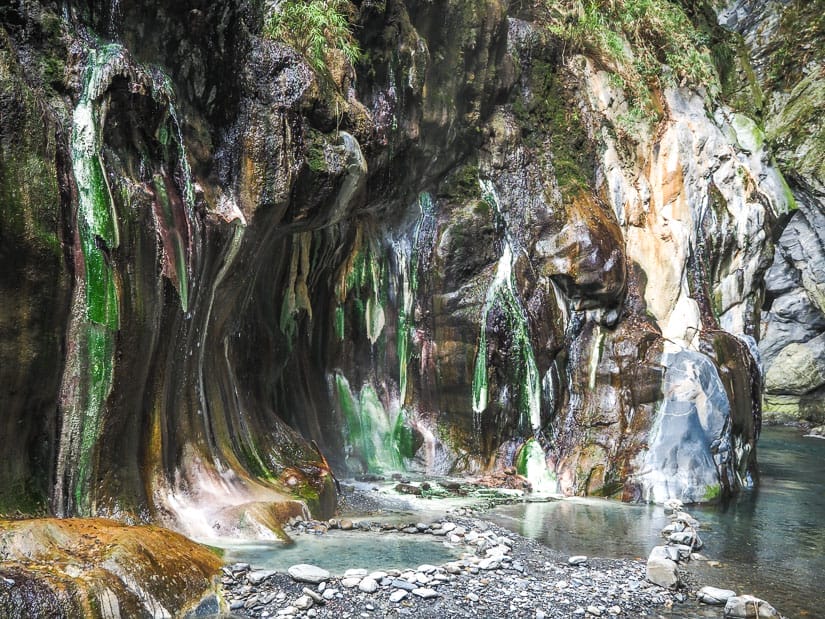 Two shallow hot spring pools formed by rocks at the base of a cliff that is covered in orange, green, and white mineral deposits