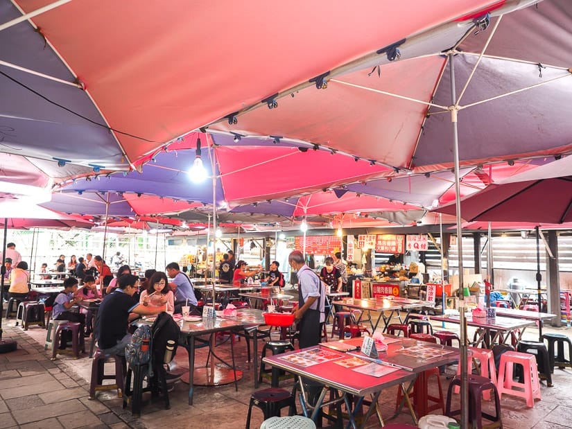 A large food court area with pink and purple canopies above and many people eating