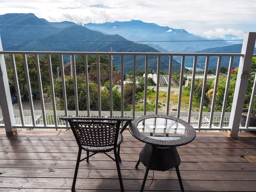 An empty seat and small table on a wooden balcony shot from behind, looking out at a rural scene and mountainous view