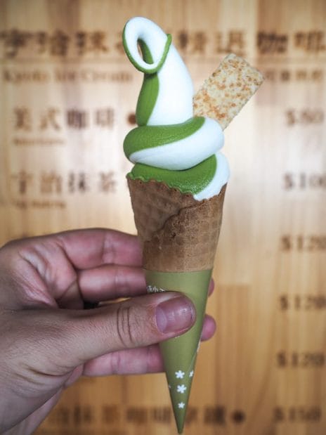 A hand holding up a soft serve ice cream waffle cone, the ice cream is half white half green matcha color, with one cookie on top, and wooden sign behind