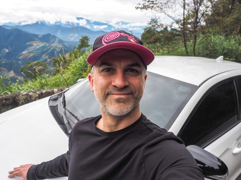 Nick Kembel taking a selfie while wearing black and pink cap, black long sleeve short, and posing on the front of a white car, with mountain scenery in background