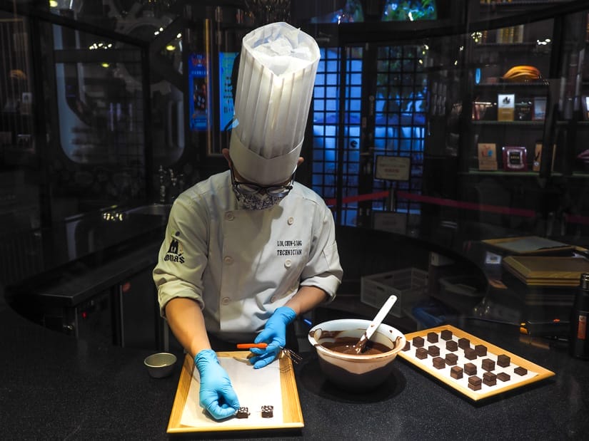 A Taiwanese chef wearing white scrubs and tall white chefs hat carefully making some chocolates on a counter