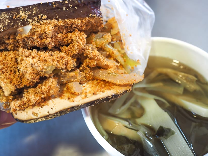 Close up of a slab of tofu stuffed with vegetables and peanut powder and a white paper bowl of bamboo soup