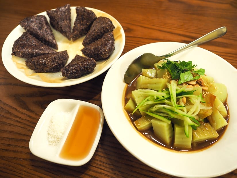 Two plates of Yunnan food on a wooden table, one is pea jelly cubes with sauce, the other is triangular chunks of purple rice, plus a small dish of sugar powder and honey
