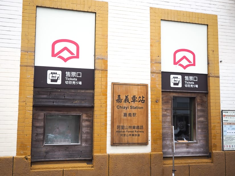 Two ticket windows on a white brick wall, with wooden frames around them and a sign in the middle that says "Alishan Forest Railway"