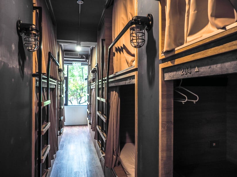 Looking down the aisle of a dorm room, with curtained capsule pods on either side and window at the end