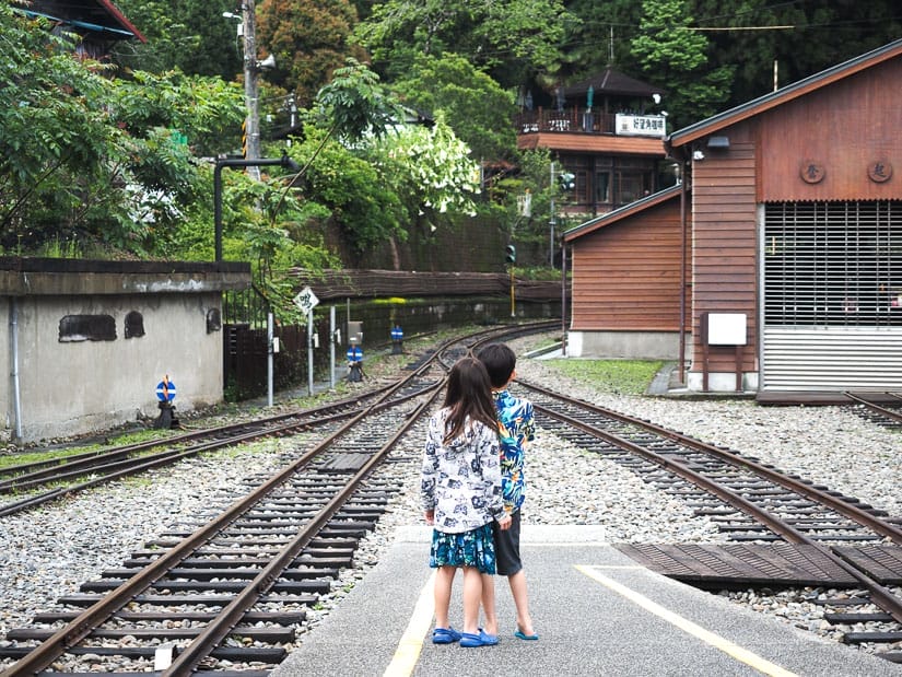 Two kids standing at the end of a cement train station platform gazing down the railway tracks