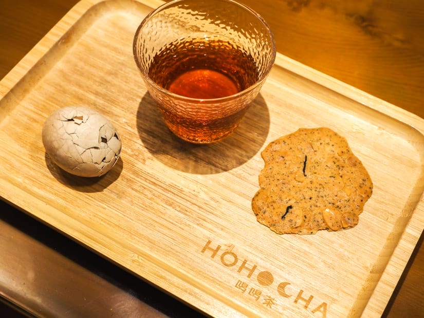 A small wooden tray with the word Hohocha, and a tea egg, glass of black tea, and tea cookie on it