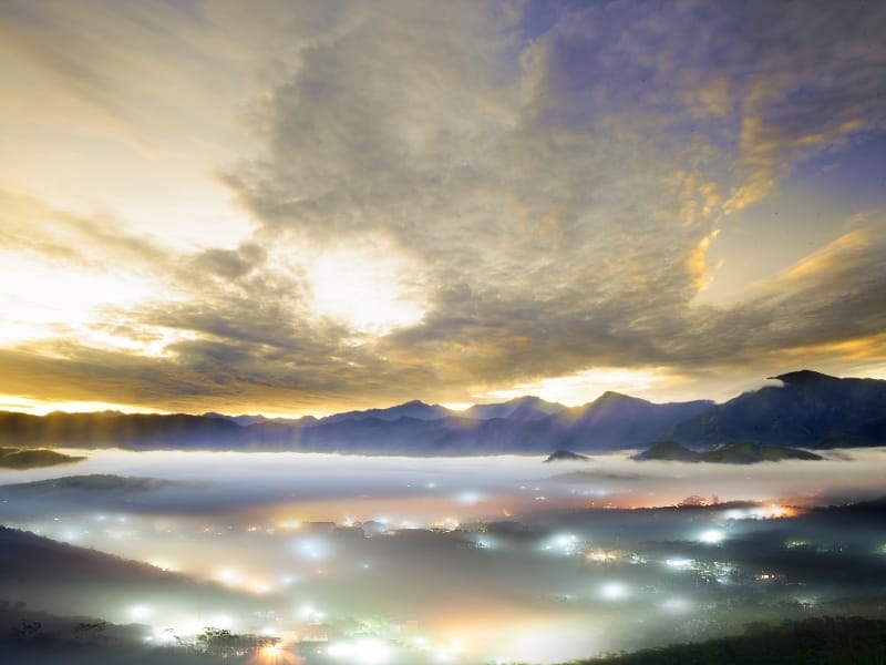 A sunrise scene, with sea of clouds covering a valley, street and house lights shining through, mountains beyond,and cloudy sky above with sunlight starting to shine through