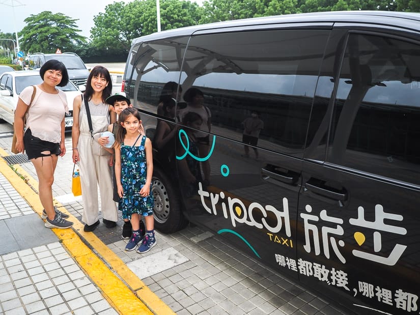 Two Taiwanese women and two kids standing beside a black van parked at the curb with the word Tripool on it