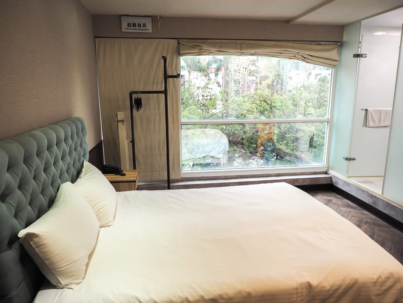 A budget hotel room with white bed, turquoise headboard, and large window with trees outside