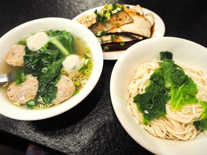 Three dishes on a table, including a soup with greens and meat balls, noodles with greens, and tofu slices with sauce