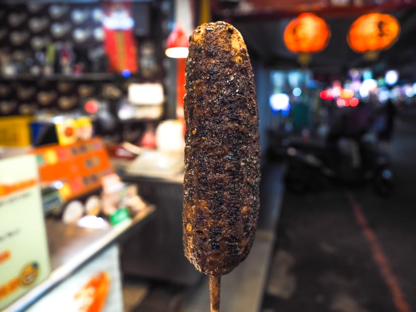 A grilled corn on the cob on a stick, upright and vertical and the center of the picture, with night market stalls in the background behind