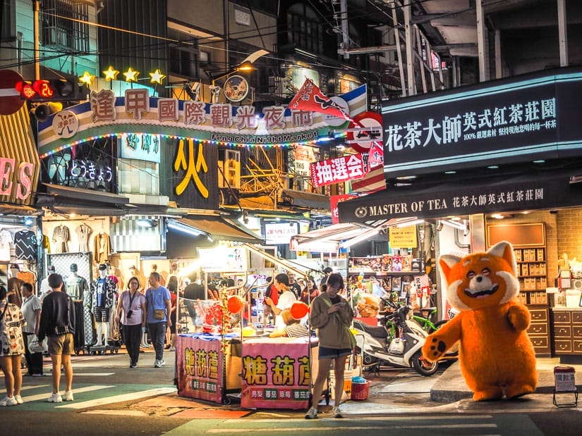 Entrance gate to Feng Chia Night Market at night, with a person dressed like a giant panda standing below it, and another person checking her phone while waiting to cross the street