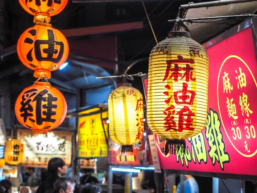 Close up of some yellow, red, and orange lanterns in a night market, with the food stall signs on the side