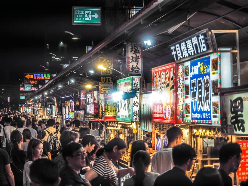 A long row of night market food stalls, with crowds of people in front of each one as they order foods
