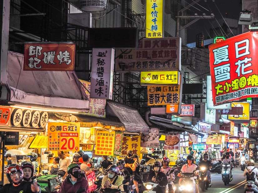 A night market scene with scooters going buy and a row of small shops
