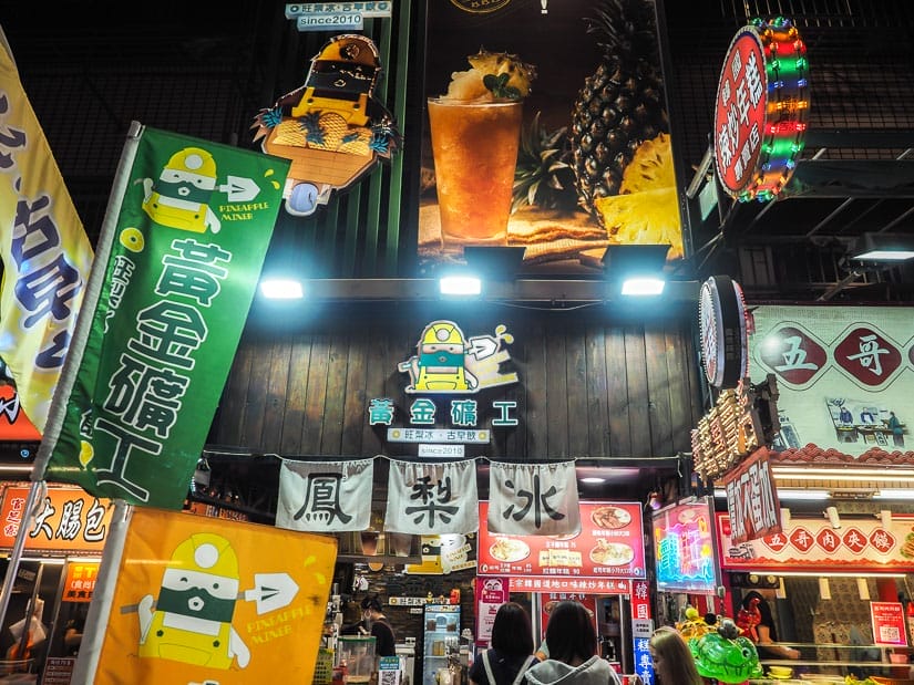 A few people shot from behind, standing in front of some night market stalls with many signs above for all the foods on offer