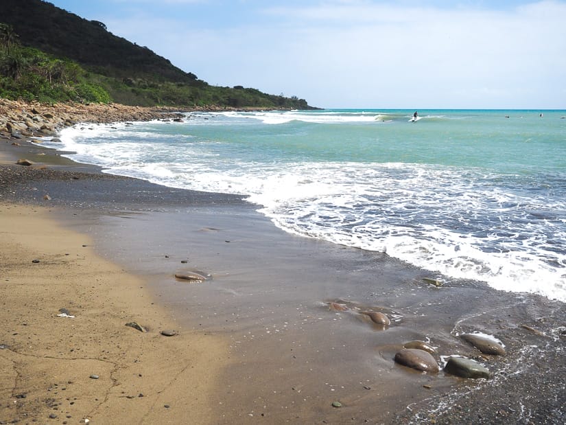 Waves rising on a brown sandy beach with some surfers visible out on the water at the back left