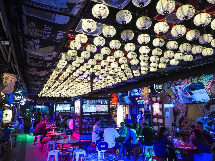 A dark, bar-like room with canopy of white lanterns above and some people sitting around eating/drinking at small tables with stools, and some neon lights lighting up the scene