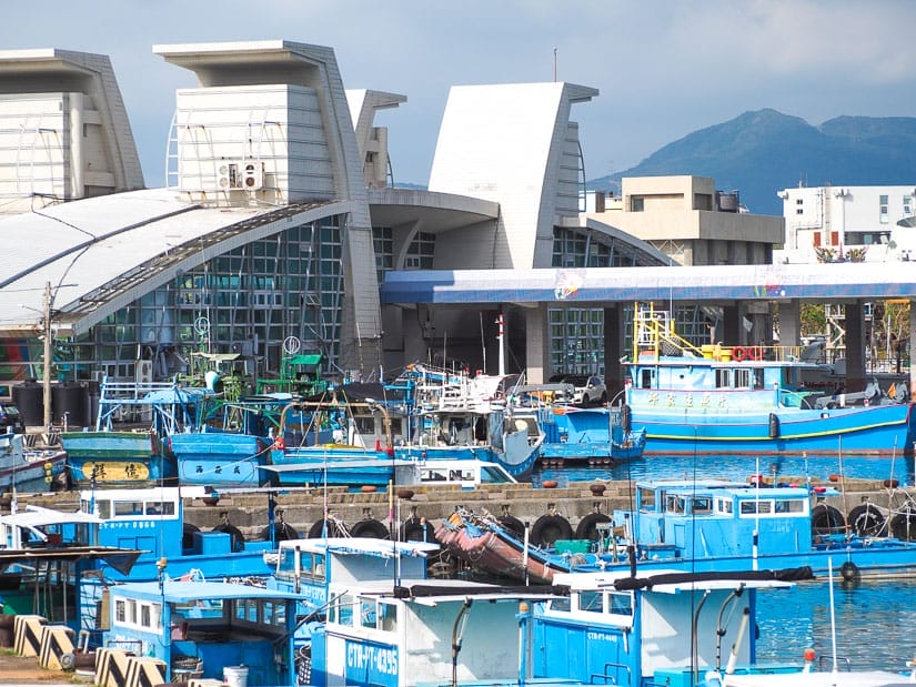 A large harbor building with habor in front of it filled with blue and white Taiwanese fishing boats