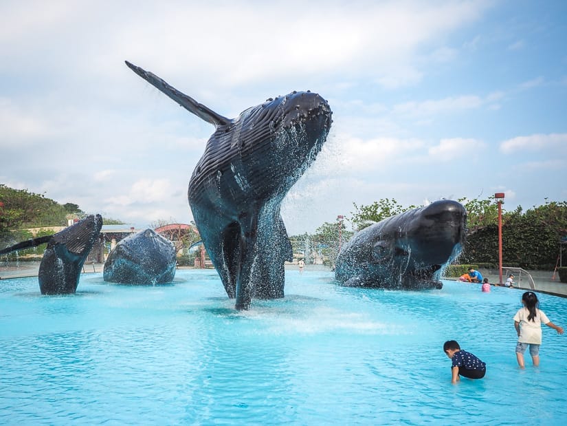 A large water fountain with giant whale statues in it and two kids playing in the water on the side