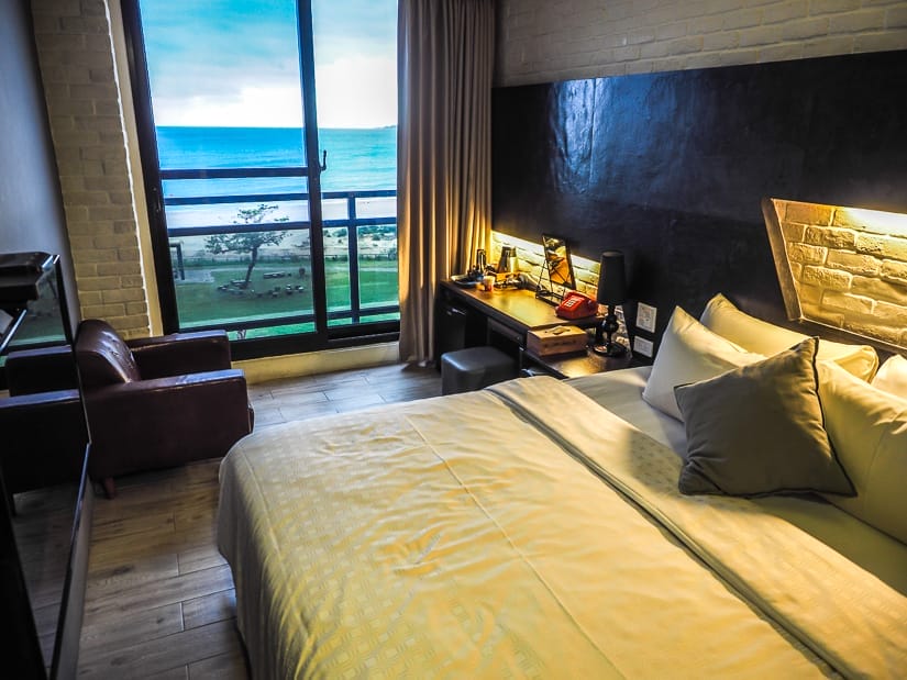 The inside of a somewhat dark hotel room with bed and glass door with view of the sea