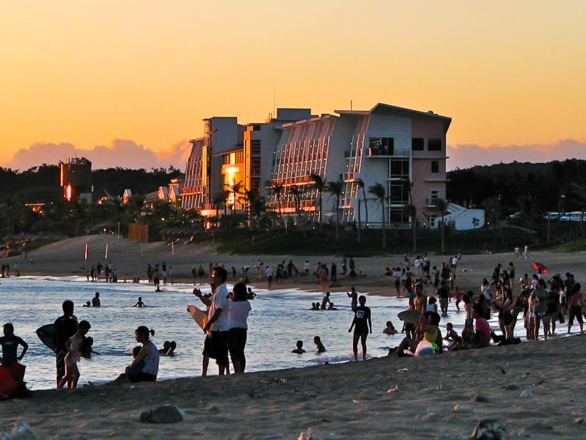 A crowd of people on a beach that's almost dark, with a beach hotel behind and orange sky from the sunset