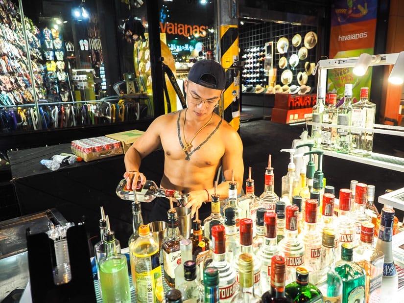 A young Taiwanese man with no short on pouring  a cocktail behind a stall full of liquor bottles