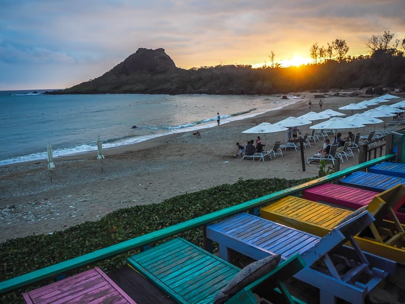 Looking down on a beach with a row of colorful bench chairs, beach in front of them, and sun setting over the coast beyond